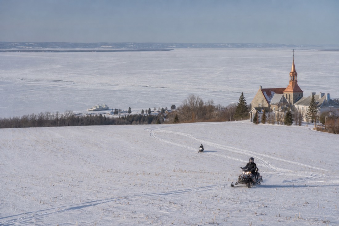 Snowmobiles crossing a snowy field on Île d'Orléans with the frozen St. Lawrence in the distance