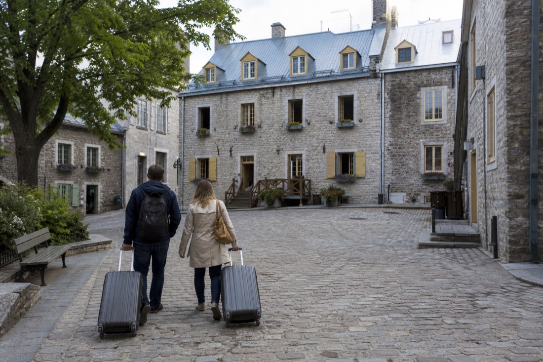 A couple with luggage crossing a cobbled square toward a small hotel in Old Quebec